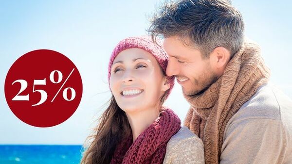 Ein Paar in Pullovern und Schals umarmt sich am Strand. Es ist sonnig. Beide lächeln glücklich. In einem roten Kreis steht 25%.
