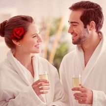 Paar in weißen Bademänteln mit Champagner-Gläsern lächelt sich an.