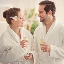 Paar in weißen Bademänteln mit Champagner-Gläsern lächelt sich an.