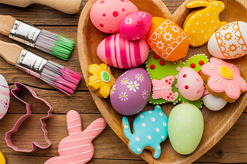 Eine herzförmige Schale mit bemalten bunten Eiern und Keksen in Hasen-Form. Daneben liegen Pinsel und Ausstechformen für Kekse in Hasen-Form.