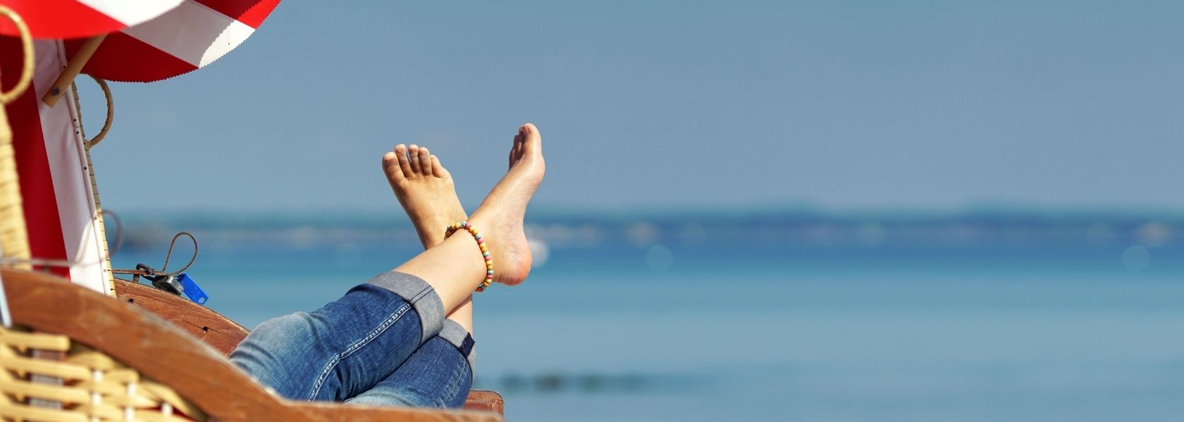 Ein Foto von einem Strandkorb am Meer, aus dem zwei Beine und Füße ohne Schuhe herausgestreckt werden. Der Himmel und das Meer sind strahlend blau.