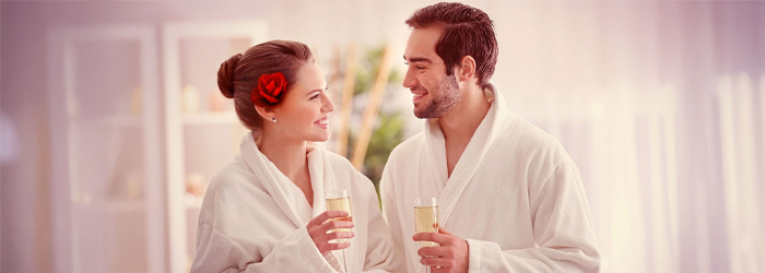 Paar in weißen Bademänteln mit Champagner-Gläsern lächelt sich in einem Spa-ähnlichen Raum an. Rote Rose im Haar der Frau.