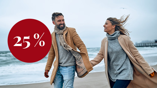 Paar in Mänteln, Pullovern, Handschuhen und Schals halten Händchen bei einem Spaziergang entlang eines Strands. Es ist stürmisch. Das Paar lächelt einander an. In einem roten Kreis steht 25%.